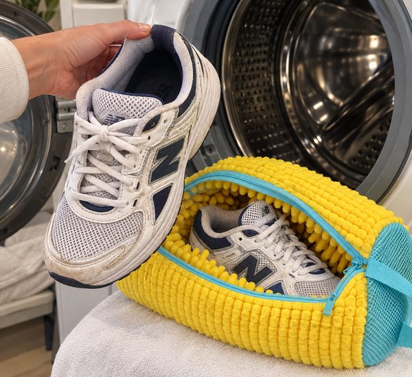 A hand holding a dirty sneaker beside the open yellow FreshKicks bag with the matching sneaker already inside, washing machine behind